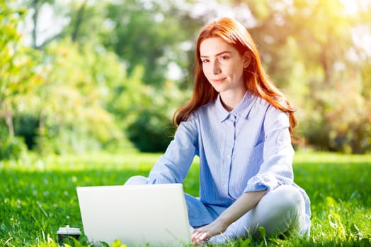 Young redhead woman using laptop computer on green grass in park. Beautiful girl in casual wear working with computer outdoors at sunny summer day. Freelance, blogging or remote work concept.