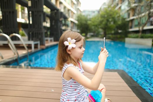 kid girl uses tablet sitting close to blue swimming pool wearing flower and hold toy. concept of new technologies travel, recreation or holiday family time on sunny day