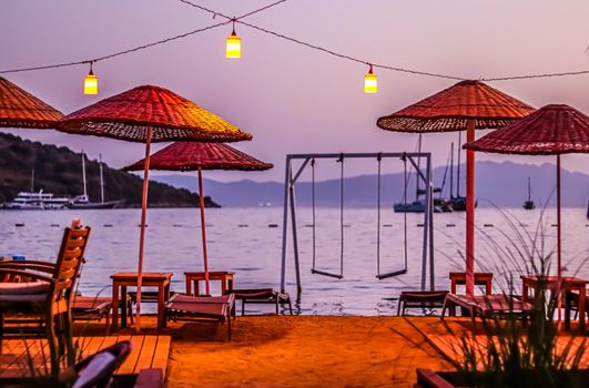 Beach umbrellas, sun loungers and swings under red lanterns against the backdrop of the sea, islands, mountains and yachts at sunset. Summer vacation concept