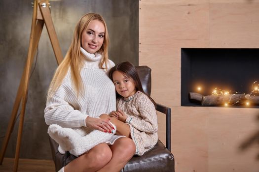 Beautiful mother woman in white sweater hugs her little daughter. Cheerful playful mood and love inside the family. Cozy home environment.