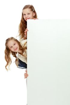 Two cute little girls look out from behind a white advertising banner. On the banner you can make any advertising inscription or put a picture or logo. It is isolated on a white background.