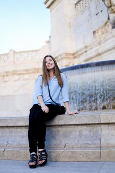 Young woman sitting near Trevi fountain, wearing blue shirt. Concept of traveling to Italy, Rome and youth.