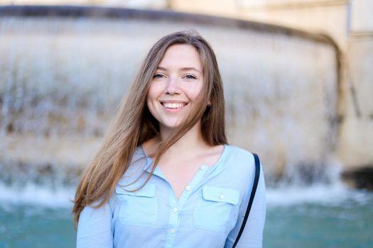 Portrait of young woman sitting near fountain and smiling, wearing blue shirt. Concept of tourism and urban resting.