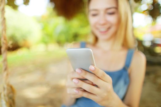 Focus on smartphone hold by young blonde girl riding swing and smiling, Thailand. Concept of modern technology, internet and summer vacations in tropics.