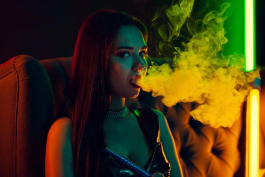 Close-up shot of a wonderful brunette lady with a perfect make-up, in a sparkling necklace and a leather dress smoking a hookah exhaling a smoke sitting on an arm-chair at a luxury night club. Concept of pleasant spending time. Dark background with colorful neon backlights.