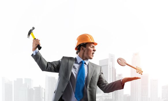 Furious businessman going to crash with hammer steel key. Young handsome contractor in business suit and safety helmet standing on cityscape background. Financial crisis and bankruptcy in real estate