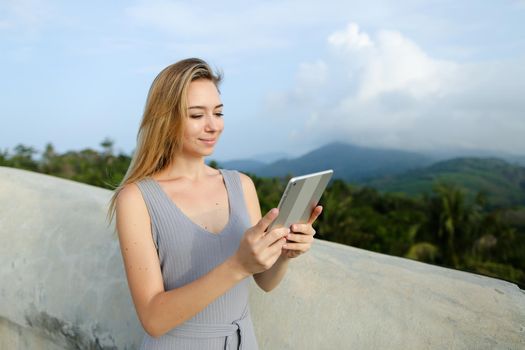 Young blonde woman using tablet in mountains background, wearing grey shirt. Concept of modern technology and nature.