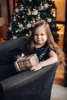 Stock photo portrait of a pretty little girl with long wavy fair hair wearing sparkling top, sitting in cozy arm-chair with wrapped Christmas present.