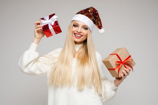 Stock photo of attractive young blonde Caucasian girl in sparkling Santa hat and white sweater holding two different beautifully wrapped Christmas gifts in hands. Smiling beauty with two Christmas presents.