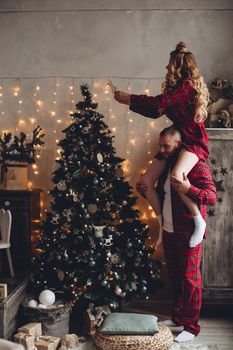 Loving couple in red checked pyjamas decorating Christmas tree. Woman sitting on man’s shoulders.