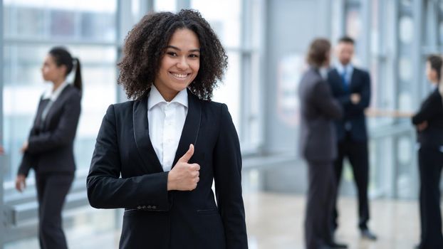 Close up portrait of young mixed race business woman standing with thumb up in front of colleagues in business building