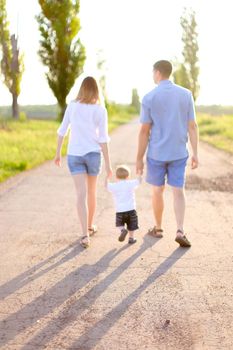 Back view of young mother and father walking on road with little baby, sunshine weather. Concept of parents and children, resting on nature.