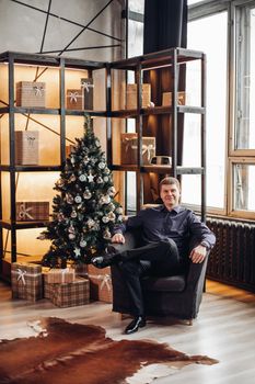 Handsome smiling man sitting relaxed in arm-chair and looking at camera. Christmas tree and shelves with wrapped Christmas gifts.