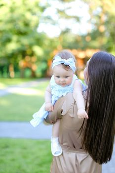 Back view of brunette young mother holding little daughter in garden. Concept of motherhood and resting on open air.