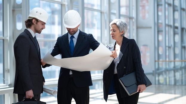 Multi ethnic business architect people in safety helmets discussing blueprint standing in the lobby at office