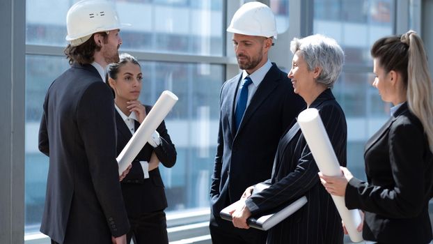 Multi ethnic business architect people in safety helmets discussing blueprint standing in the lobby at office