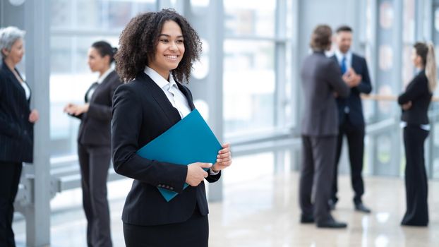 Close up portrait of young mixed race business woman standing with document folder in front of colleagues in business building