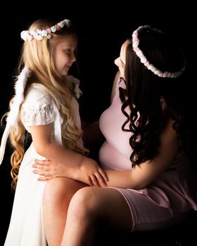 A little girl in the image of an angel hugs the belly of her eldest pregnant sister. The concept of family happiness, the birth of a child.