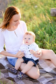 Young happy woman sitting with little baby on plaid, grass on background. Concept of picnic, motherhood and nature.