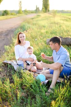 Young american mother and father sittling on grass with little baby and blowing bubbles. Concept of picnic and children, parenthood and leisure time.