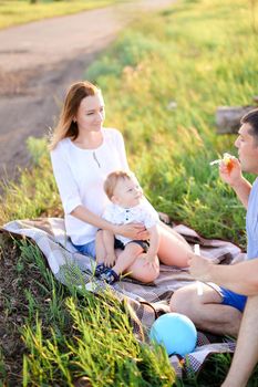 Young mother and father sittling on grass with little baby and blowing bubbles. Concept of picnic and children, parenthood and leisure time.