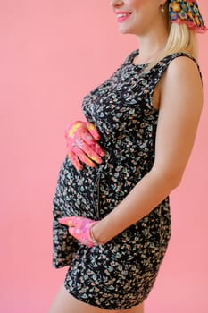 Young pregnant female person holding belly and wearing gloves in pink monophonic background. Concept of expactant female person and phot session at studio.