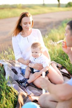 Young happy caucasian mother and father sittling on grass with little baby and blowing bubbles. Concept of picnic and children, parenthood and leisure time.