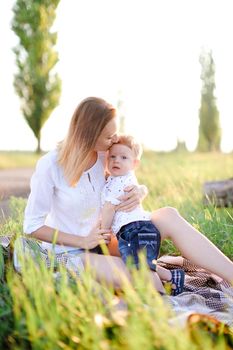 Young happy woman kissing little baby on plaid, grass on background. Concept of picnic, motherhood and nature.