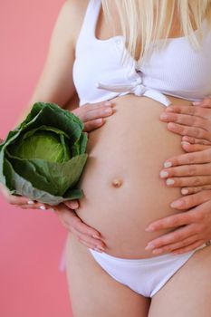 Closeup pregnant woman in underwear holding belly and keeping cabbage in pink monophonic background. Concept of expactant photo session.