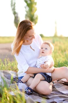 Young caucasian woman sitting with little baby on plaid, grass on background. Concept of picnic, motherhood and nature.