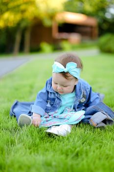 Little caucasian female baby sitting on green grass and wearing jeans jacket. Concept of childhood and resting in garden, nature background.