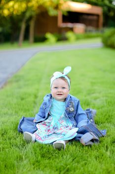 Little female baby sitting on green grass and wearing jeans jacket. Concept of childhood and resting in garden, nature background.