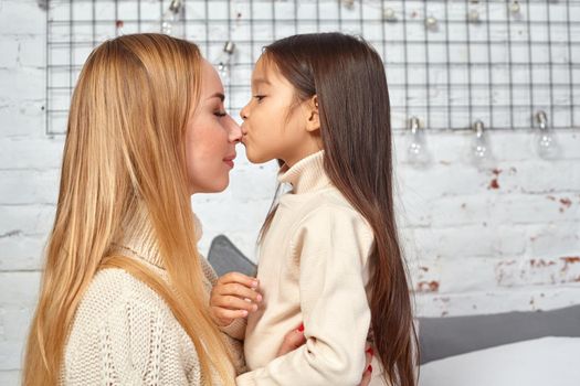 Beautiful young woman and her charming little daughter in white sweaters and jeans lying on the bed at home, are hugging.