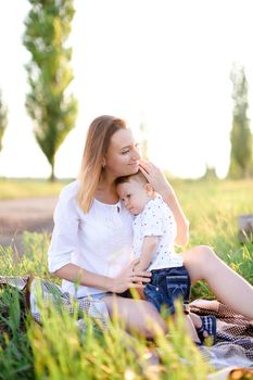 Young european woman sitting with little baby on plaid, grass on background. Concept of picnic, motherhood and nature.