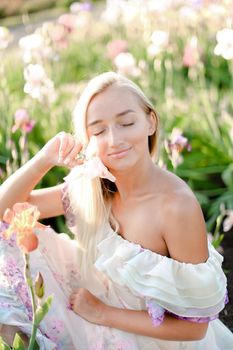 Young smiling woman sitting near irises on garden, wearing white dress. Concept of heman beauty and spring inspiration.