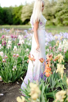 Young woman wearing white dress standing near irises on garden. Concept of human beauty and flora, spring inspiration.