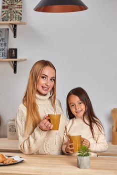 Shot of cheerful mother and daughter sit together at kitchen table, drink hot tea in morning, have pleasant friendly talk between each other. Curious girl asks something in mum during coffee break.