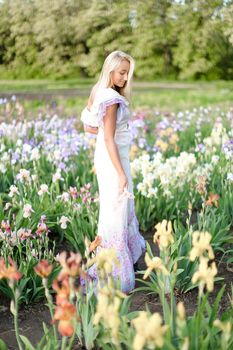 Young european woman wearing white dress standing near irises on garden. Concept of human beauty and flora, spring inspiration.