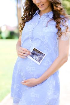 Happy young pregnant girl wearing blue dress holding belly with ultrasound photo. Concept of expectant female person and motherhood.