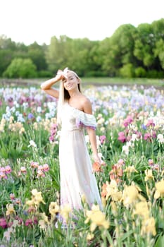 Young woman wearing white dress standing near irises on garden. Concept of human beauty and flora, spring inspiration.