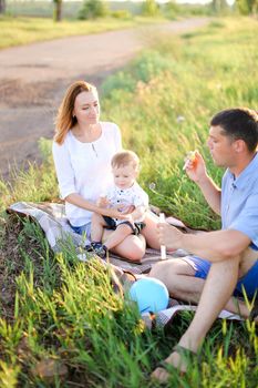 Young gladden mother and father sittling on grass with little baby and blowing bubbles. Concept of picnic and children, parenthood and leisure time.