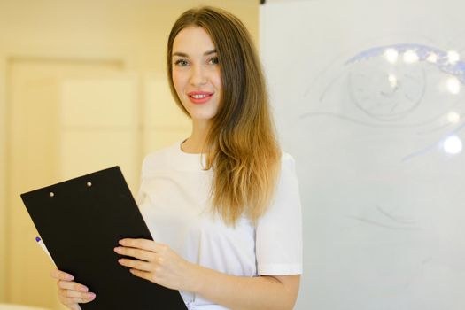 Caucasian cosmetologist standing with black folder near placard with drawn eye at cosmetology cabinet. Concept of qualitative medical worker, beauty salon and permanent makeup.