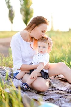 Young woman kissing little baby and sitting on plaid, grass on background. Concept of picnic, motherhood and nature.