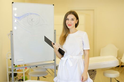 Young female cosmetologist standing with black folder near placard with drawn eye at cosmetology cabinet. Concept of qualitative medical worker, beauty salon and permanent makeup.