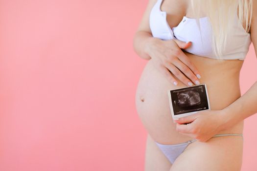 Young happy pregnant woman wearing underwear and holding belly with ultrasound in pink monophonic background. Concept of expectant photo session at studio.