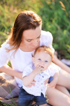 Young tender woman kissing little baby on plaid, grass on background. Concept of picnic, motherhood and nature.