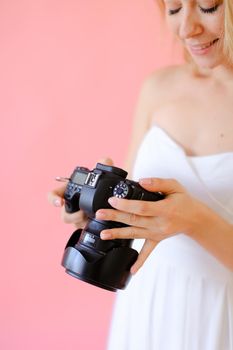 Closeup young blonde gladden female photographer with black camera in pink monophonic background. Concept of modern profession and photo session.