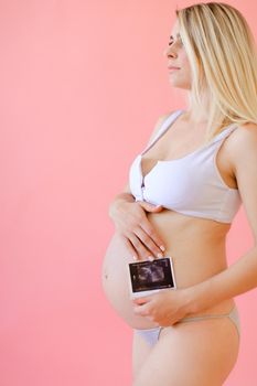 Young happy pregnant woman wearing underwear and holding belly with ultrasound in pink monophonic background. Concept of expectant photo session at studio.