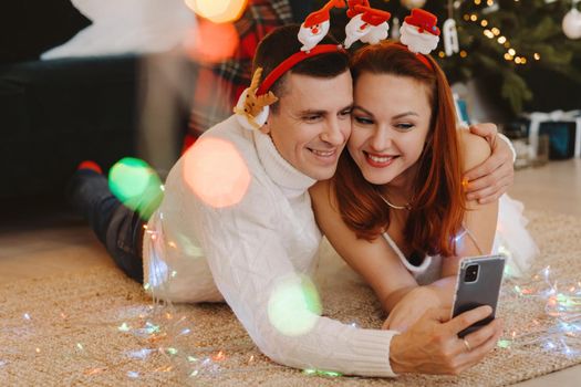 a happy married couple is lying on the floor at home near the Christmas tree and taking pictures of themselves.