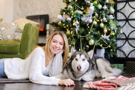 Young smiling girl lying on floor with adult malamute near Christmas tree. Concept of celebrating New Year, winter vations and pets.